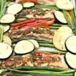 a baking sheet with 4 salmon fillets seasoned with herbs and sliced zucchini and squash and asparagus