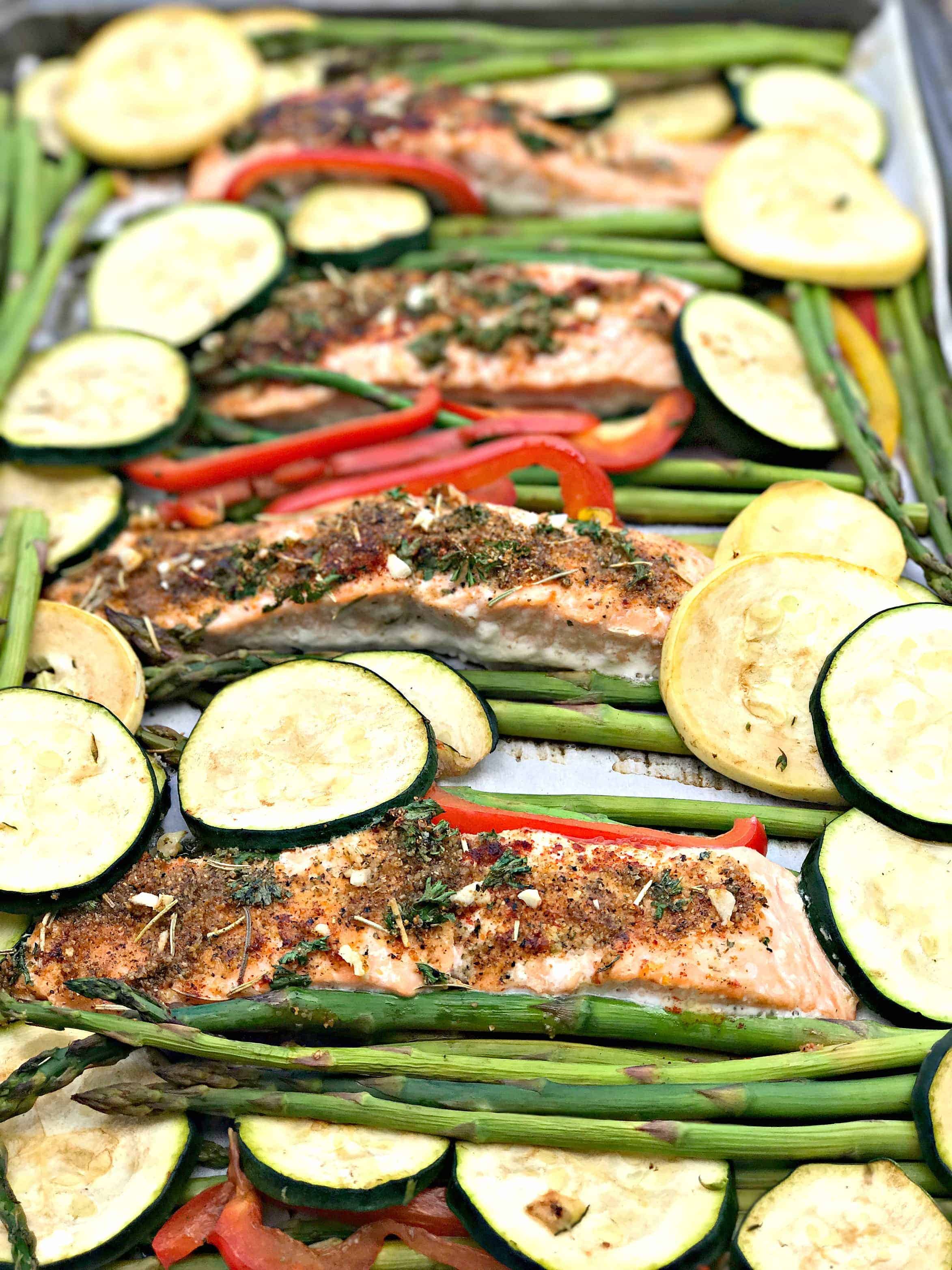 a baking sheet with 4 salmon fillets seasoned with herbs and sliced zucchini and squash and asparagus