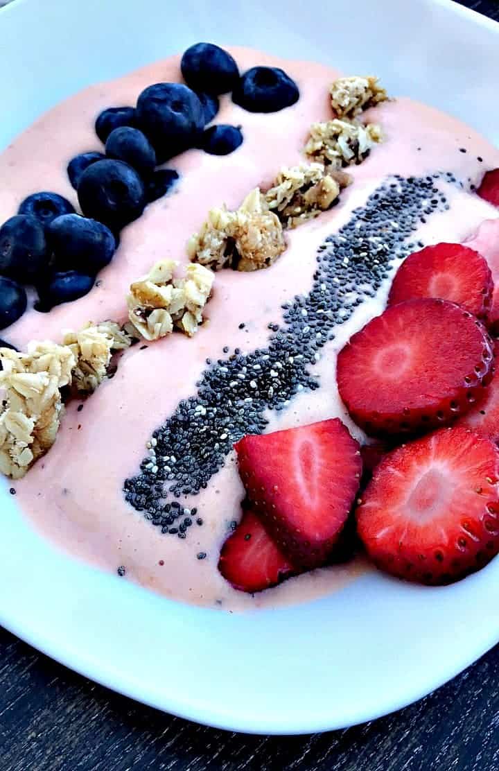 a white bowl filled with strawberry protein smoothie bowl and topped with blueberries, granola, chia seeds, and sliced strawberries