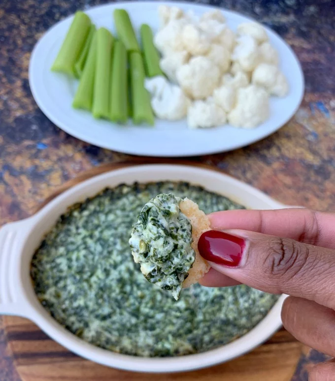 person holding cheese crisp with keto low carb spinach dip in a bowl with vegetables on a white plate