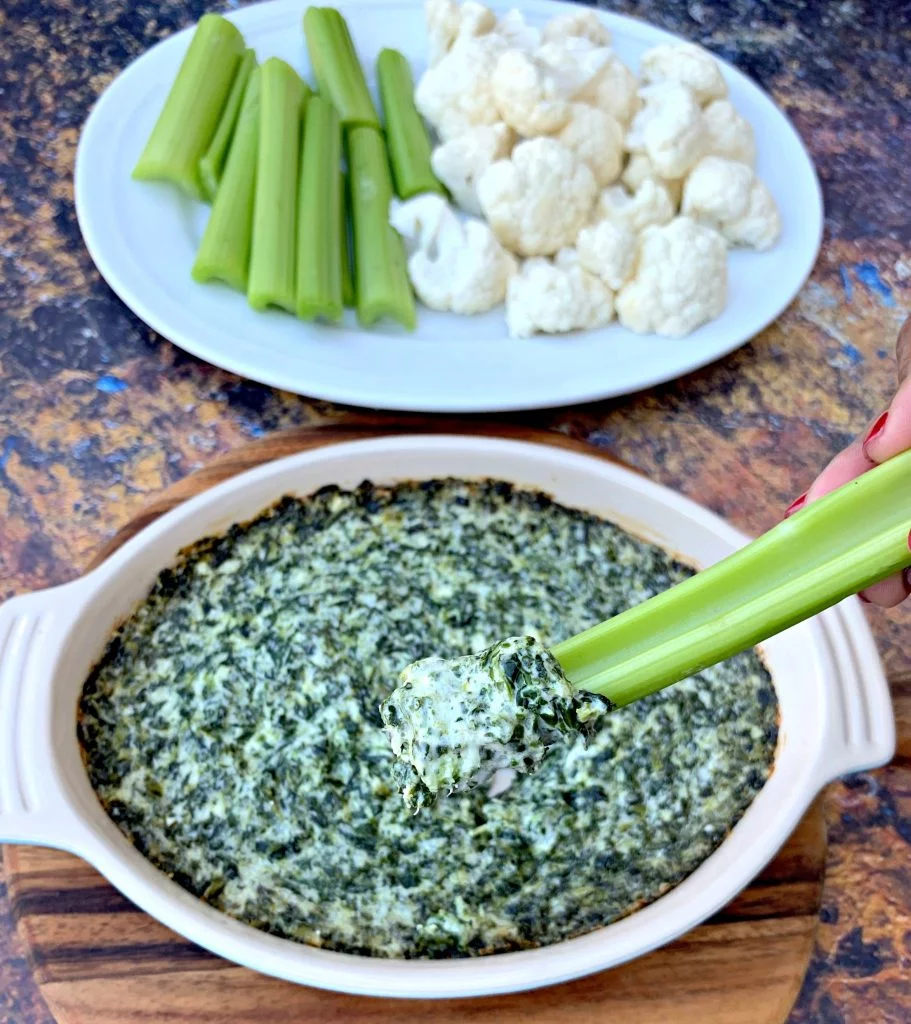 person holding celery with keto low carb spinach dip in a bowl with vegetables on a white plate