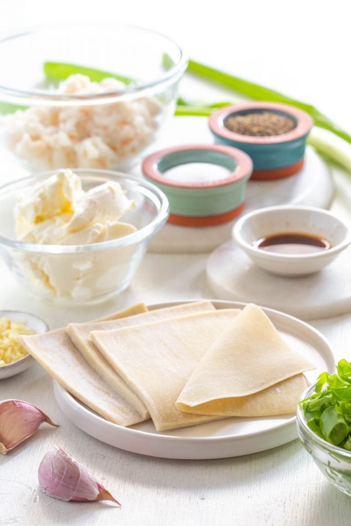 wonton wrappers, cream cheese, lump crab, and green onions in separate bowls on a flat surface