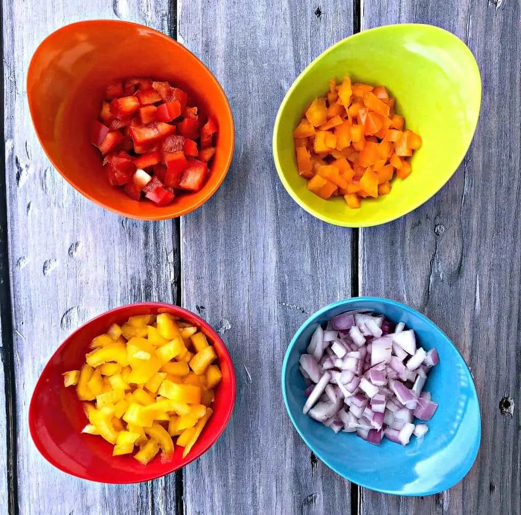 red, orange, and yellow peppers diced in multi colored bowls and red onions in a blue bowl