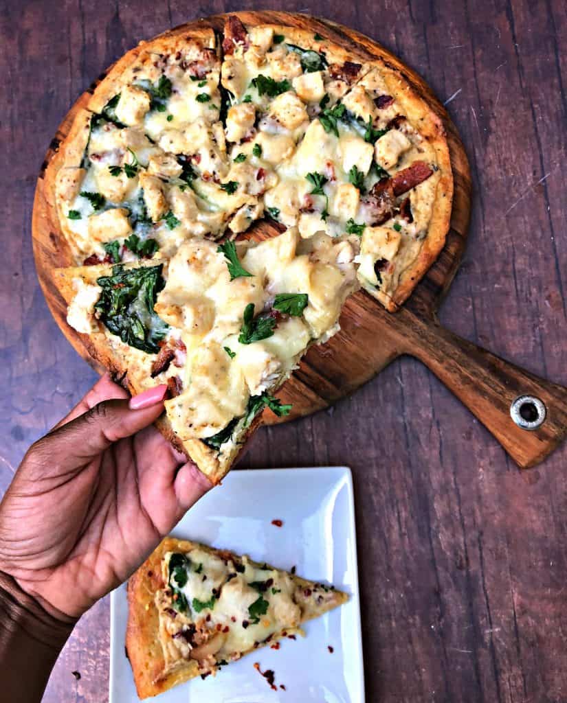 person holding slice of keto fathead alfredo bacon chicken pizza on a pan