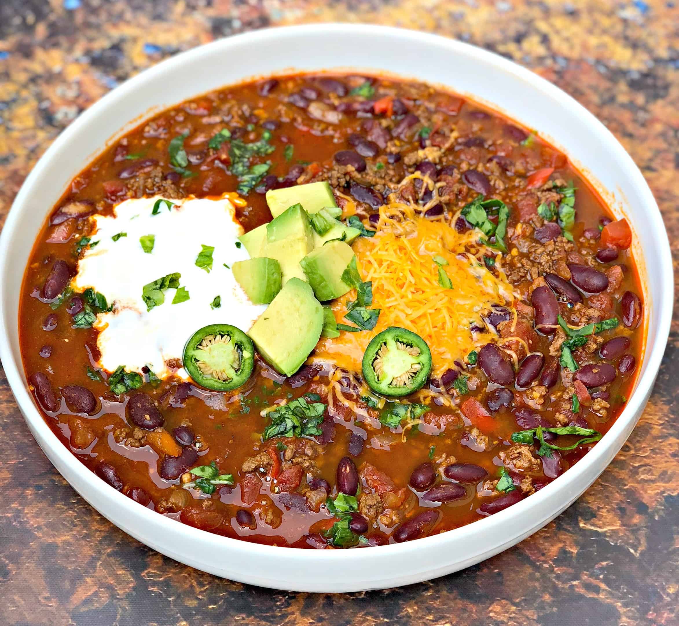 instant pot chili with shredded cheese, avocado, sour cream and jalapeno served in a white bowl