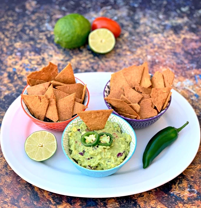 Keto Low-Carb Gluten-Free Homemade Tortilla Chips are a quick and easy recipe that uses a handful of ingredients. These crispy, crunchy chips are made with almond flour, Himalayan salt, and mozzarella and are practically no carb/carb free. You can also make crackers using the dough. The post also includes information on where to buy keto chips and what brands to look out for. #KetoRecipes #KetoTortillaChips keto tortilla chips in bowls on a plate with guacamole and vegetables