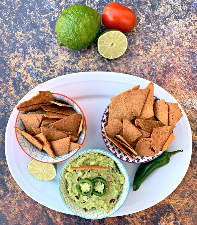 Keto Low-Carb Gluten-Free Homemade Tortilla Chips are a quick and easy recipe that uses a handful of ingredients. These crispy, crunchy chips are made with almond flour, Himalayan salt, and mozzarella and are practically no carb/carb free. You can also make crackers using the dough. The post also includes information on where to buy keto chips and what brands to look out for. #KetoRecipes #KetoTortillaChips keto tortilla chips in bowls on a plate with guacamole and vegetables