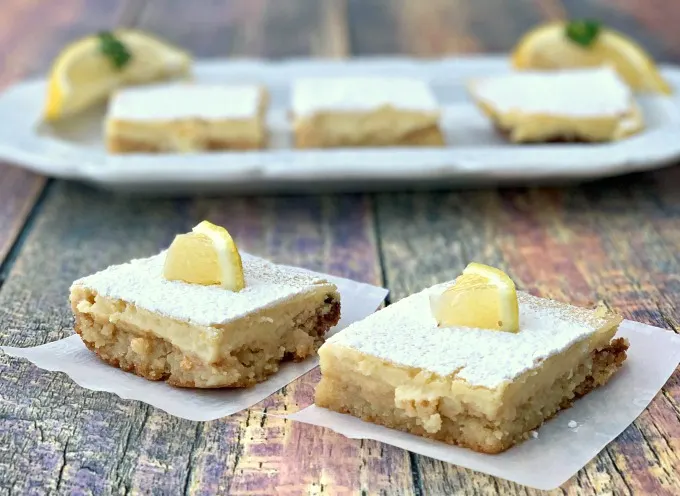 2 keto lemon bars on parchment paper with 3 lemons bars on a white plate