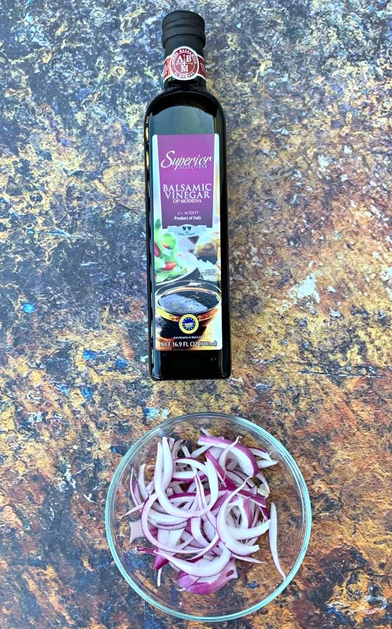 a bottle of balsamic vinegar alongside sliced red onions in a bowl