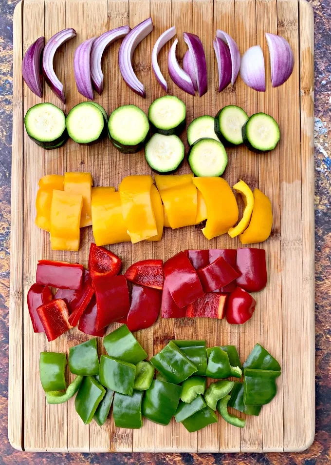 raw red peppers, green pepper, yellow peppers, zucchini, and red onions on a cutting board