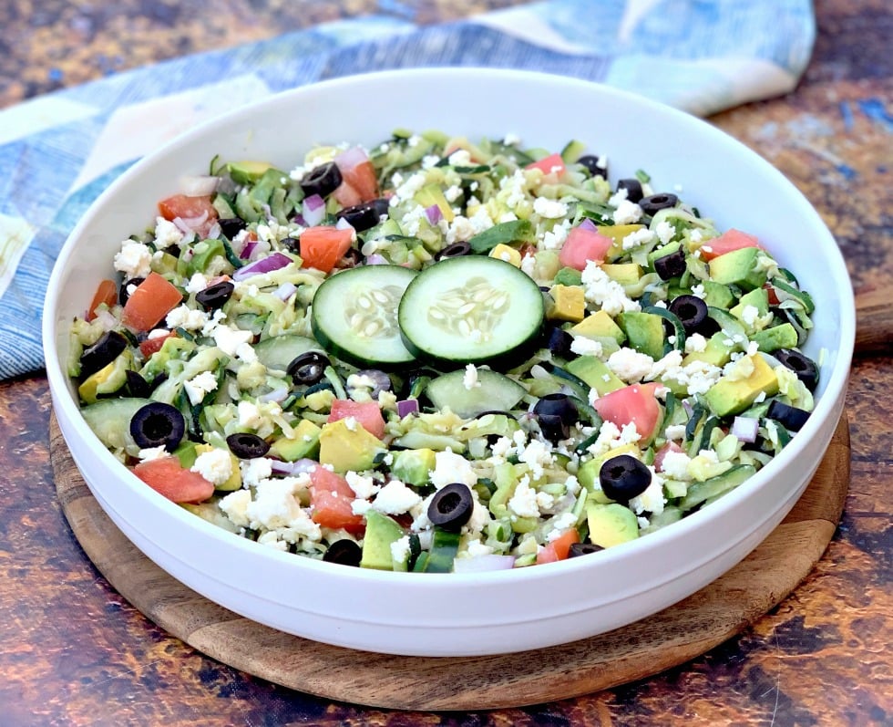 Keto Low-Carb Zucchini Noodle Pasta Salad is the best healthy recipe using raw spiralized deli veggies, cucumbers, avocado, and Green Goddess dressing. This keto-friendly, paleo pastaless salad is a spin on Greek salad. Toss with shrimp or chicken if you wish! #KetoRecipes #KetoPastaSalad keto zucchini noodles pasta salad in a white bowl with tomatoes, olives, cucumbers, feta cheese, and red onions