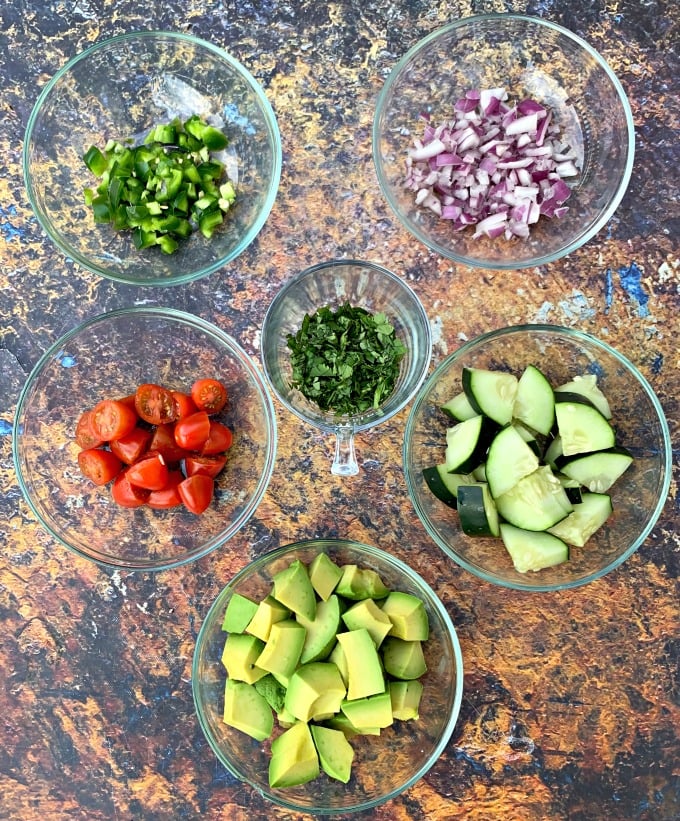 chopped jalapenos, onions, avocado, cucumbers, cilantro, and tomatoes in glass bowls on a flat surface