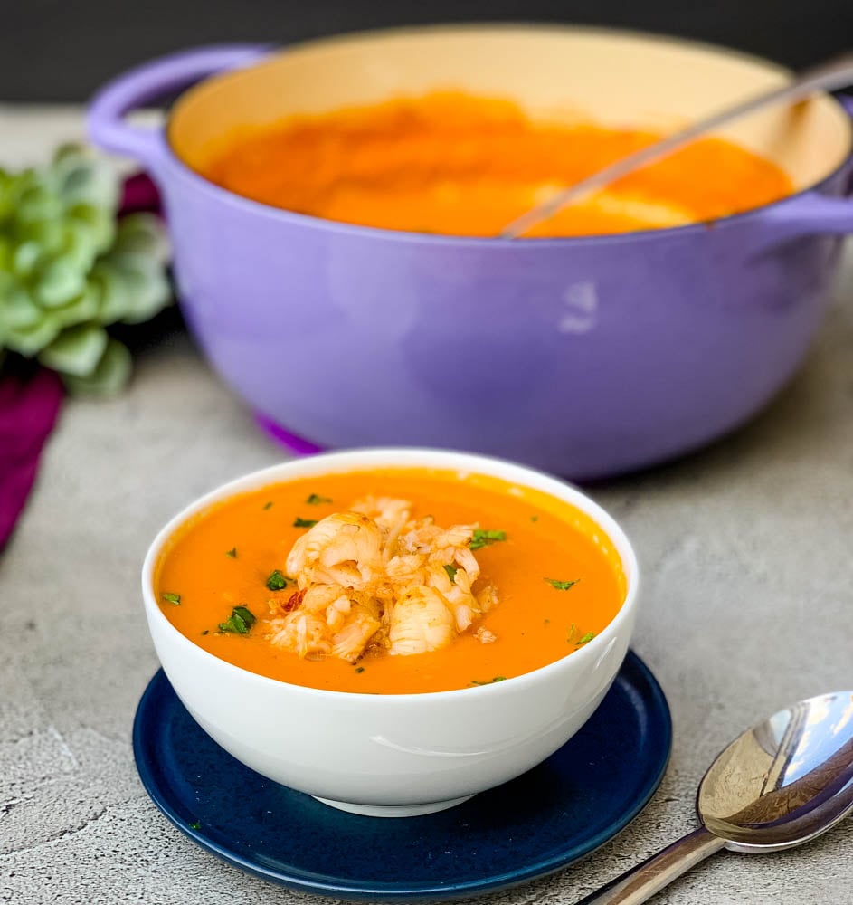 creamy homemade lobster bisque recipe in a white bowl with a purple towel and purple Dutch oven