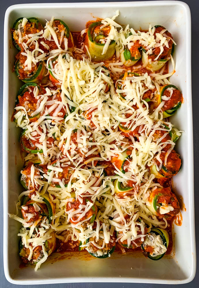 zucchini lasagna roll ups in a white baking pan with shredded cheese