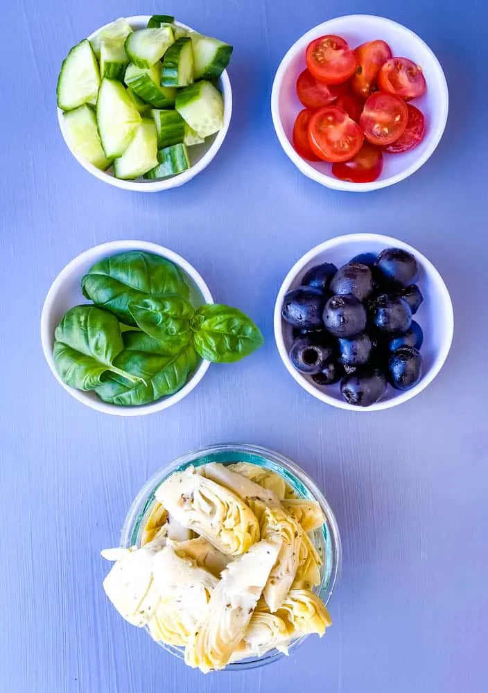 black olives, artichokes, cucumbers, and sliced cherry tomatoes in separate white bowls for antipasto salad recipe