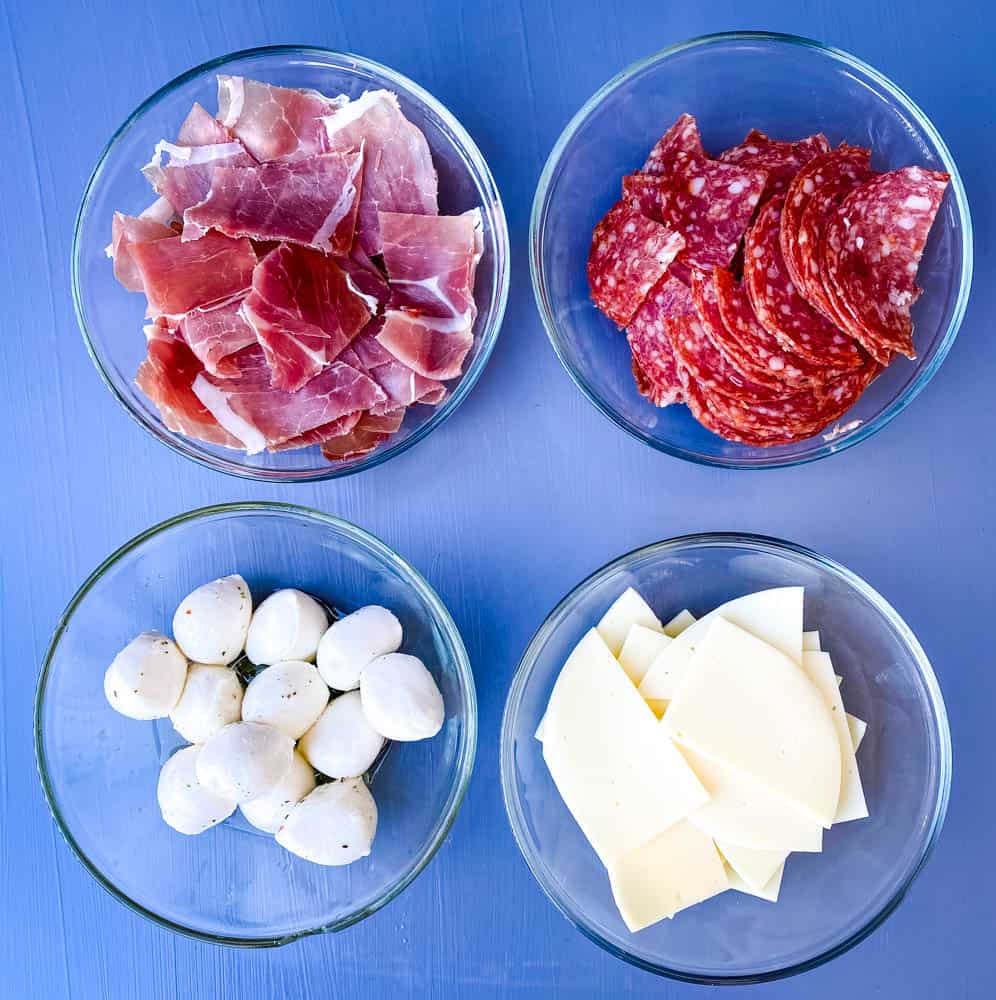 prosciutto, salami, mozzarella balls, and sliced provolone cheese in separate white bowls for antipasto salad recipe