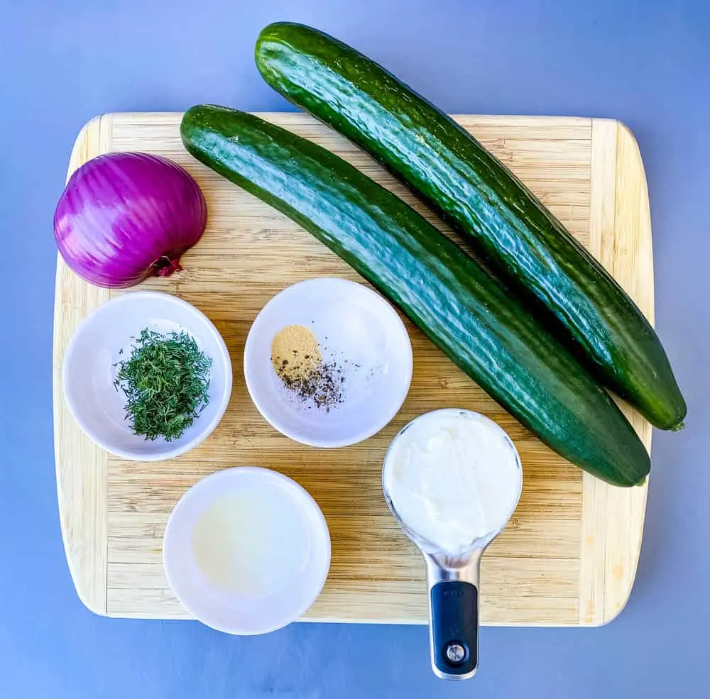 fresh cucumber and a red onion on a bamboo cutting board with a cup of Greek yogurt, fresh dill, seasoning, and lemon juice in separate bowls