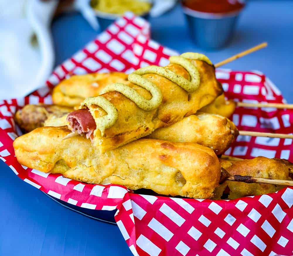 a basket of air fryer corn dogs, one corn dog with mustard and bite taken out of it