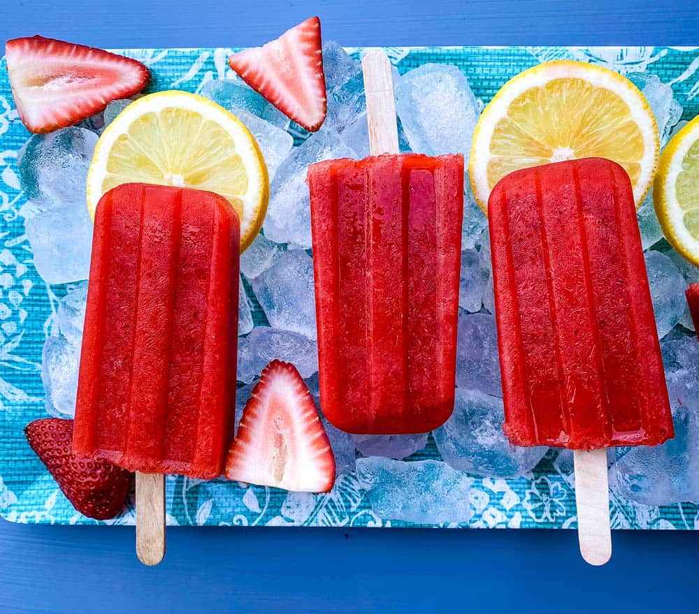 sugar free strawberry lemon popsicles on a flat surface with ice, fresh lemons and strawberries