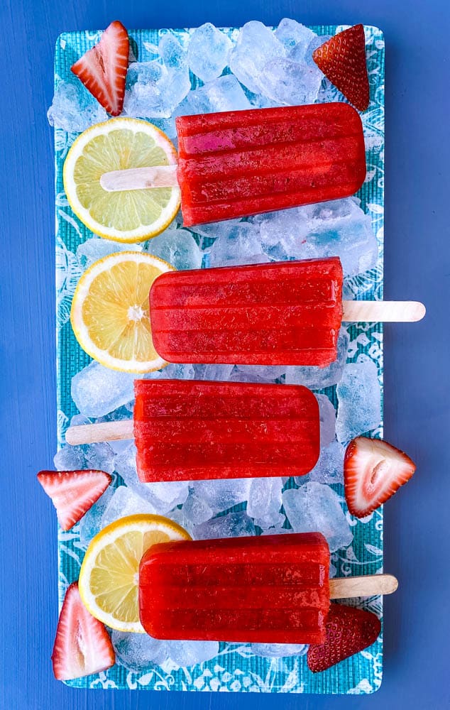 sugar free strawberry lemon popsicles on a flat surface with ice, fresh lemons and strawberries