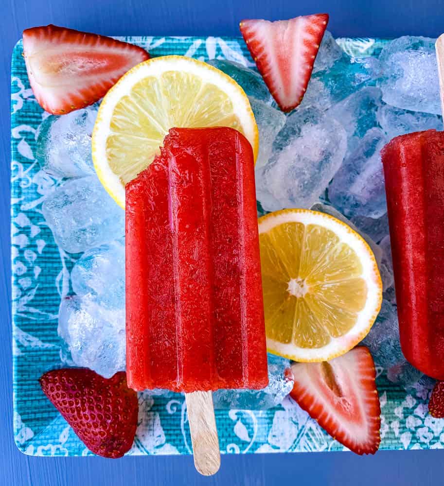 sugar free strawberry lemon popsicle with a bite removed on a flat surface with ice, fresh lemons and strawberries 