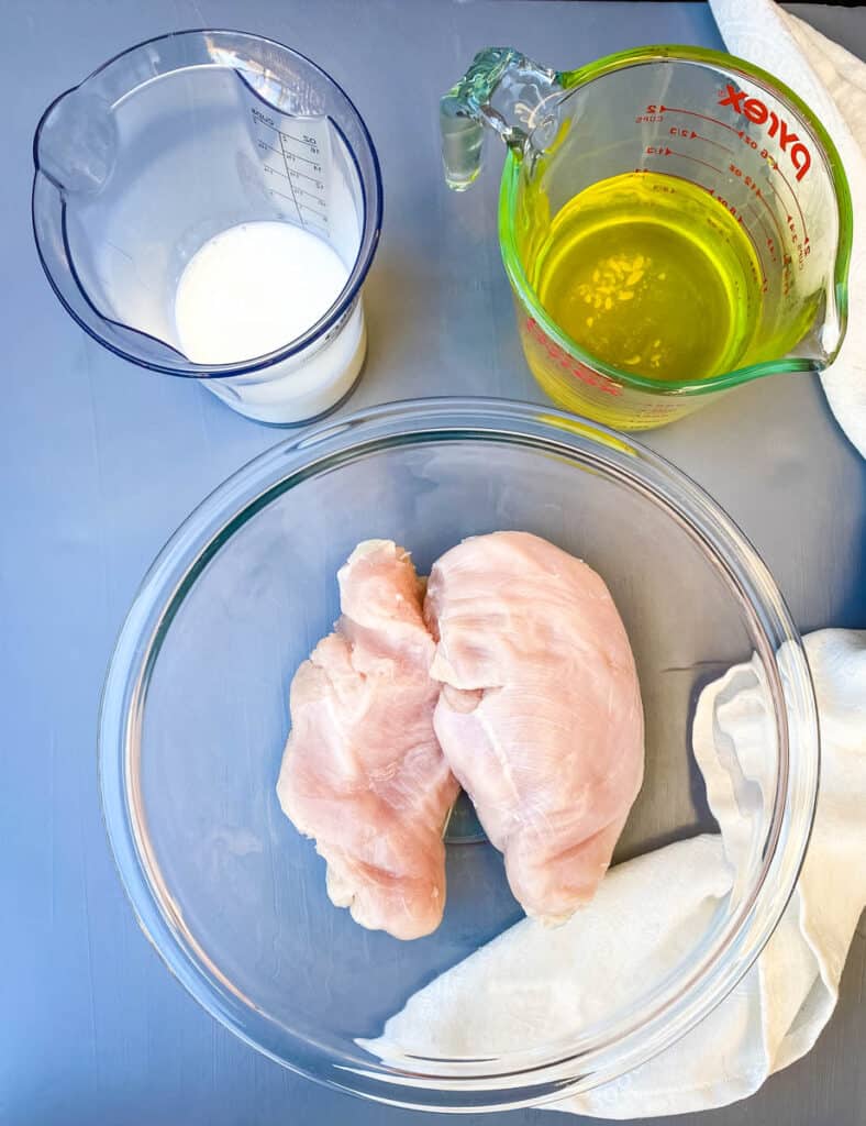 raw chicken breasts in a glass bowl, buttermilk in cup, and pickle juice in a cup