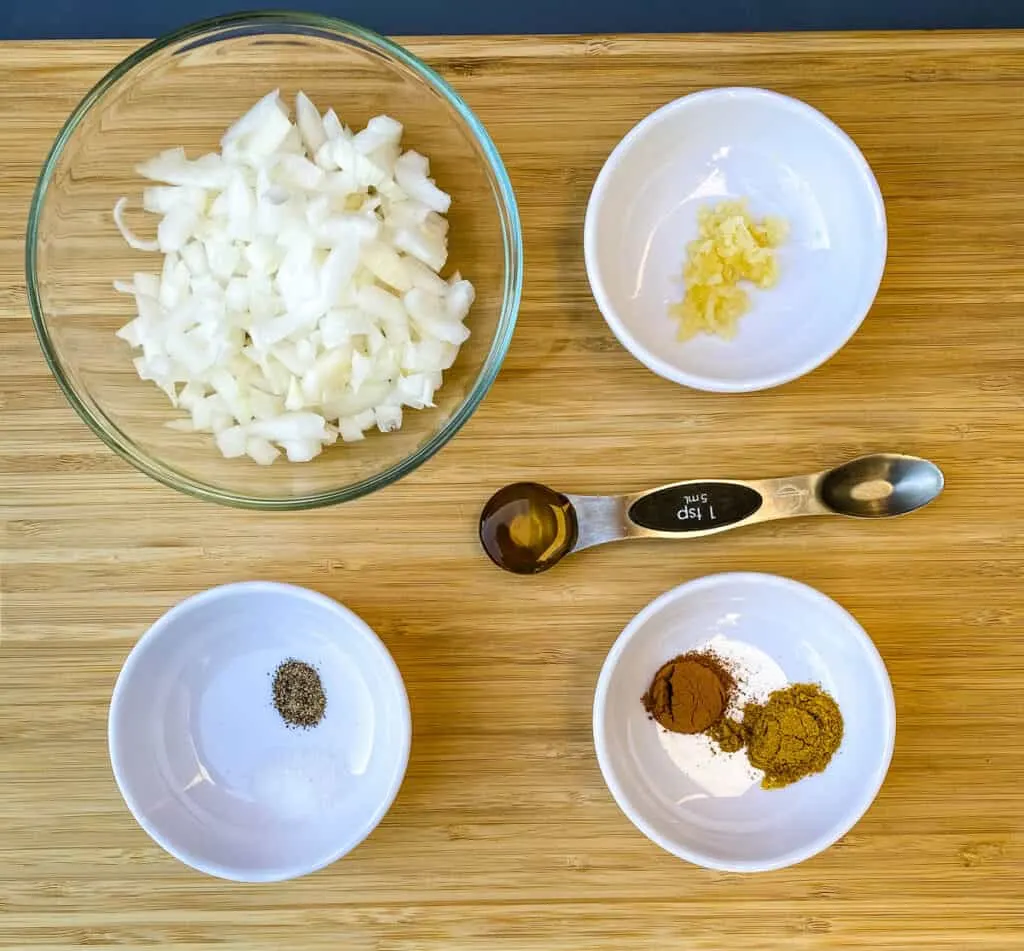 chopped onions, garlic, cinnamon, curry, salt, pepper, and honey on a wooden cutting board