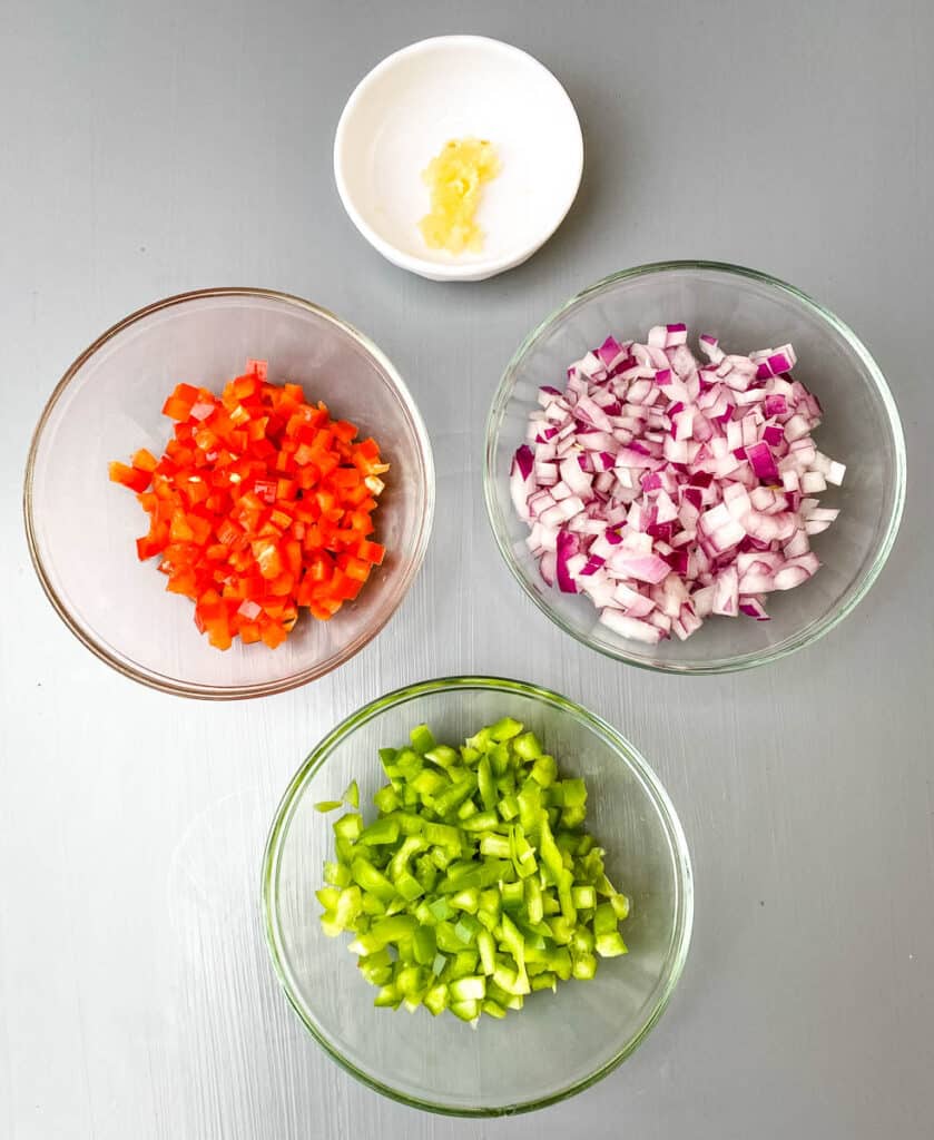 raw green peppers, red peppers, chopped onions, and garlic in separate bowls on a flat surface