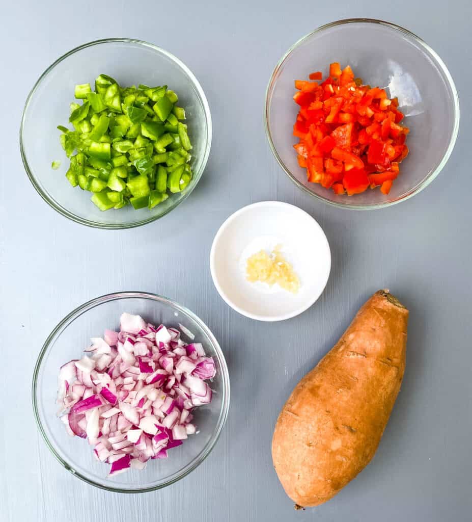 chopped green peppers, red peppers, onion, garlic, and a raw sweet potato on a flat surface