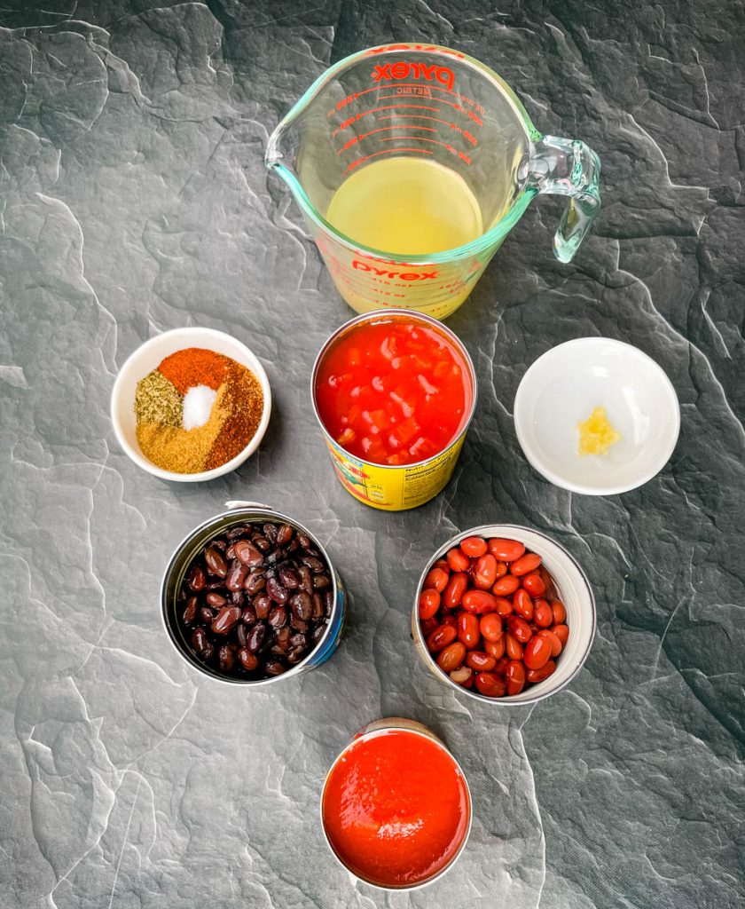 broth, black beans, kidney beans, chili seasoning, tomato sauce petite tomatoes, and garlic in separate bowls on a flat surface