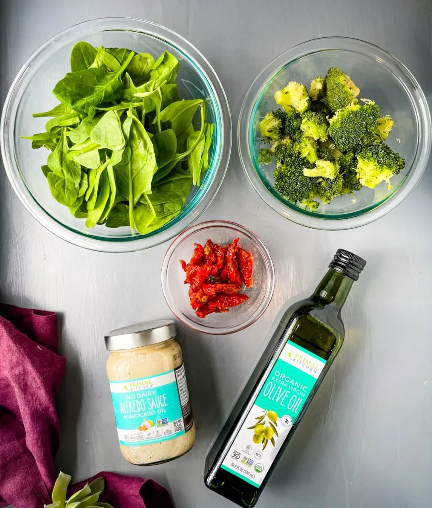 fresh spinach, fresh broccoli, sundried tomatoes, primal kitchen alfredo, and primal kitchen olive oil on a flat surface