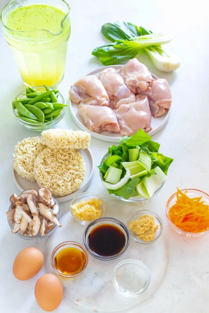 raw chicken thighs, bok choy, eggs, ramen, and vegetables in separate bowls on a counter