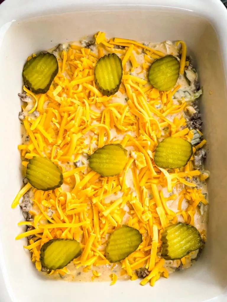 cooked ground beef in a casserole dish topped with shredded cheddar cheese and pickles