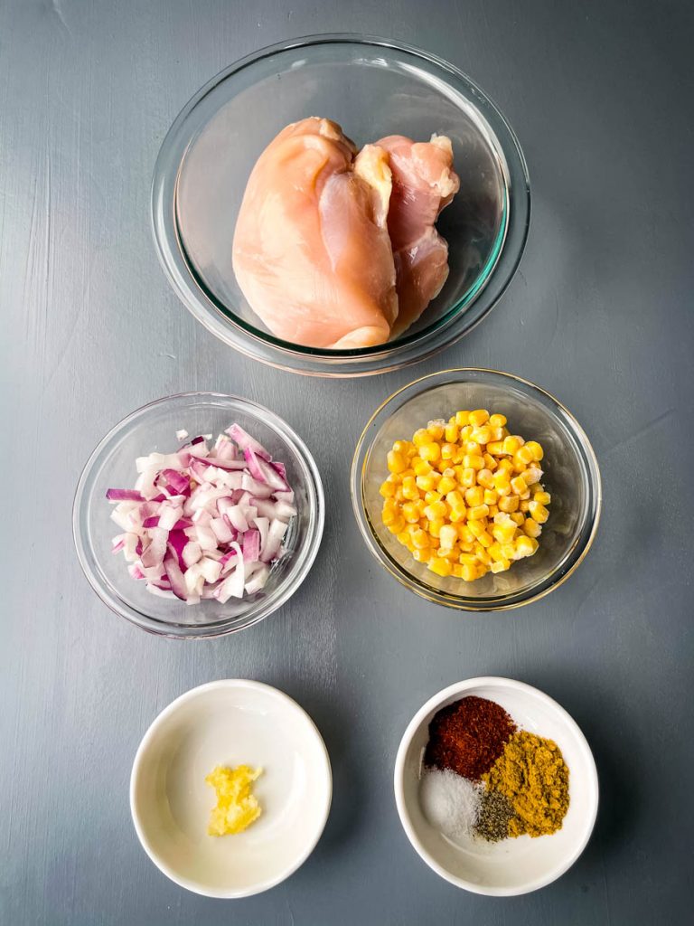 raw chicken breasts, chopped red onions, minced garlic, frozen corn, and chili seasoning in separate bowls on a flat surface