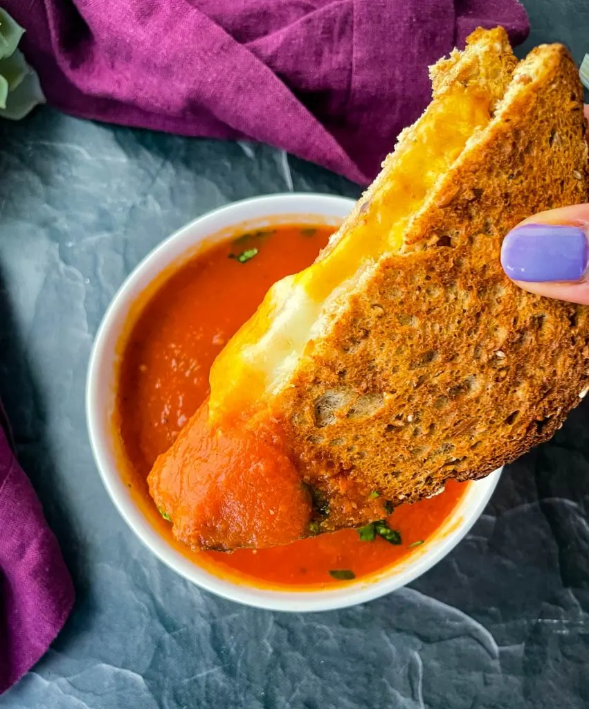 person holding a grilled cheese sandwich over tomato soup