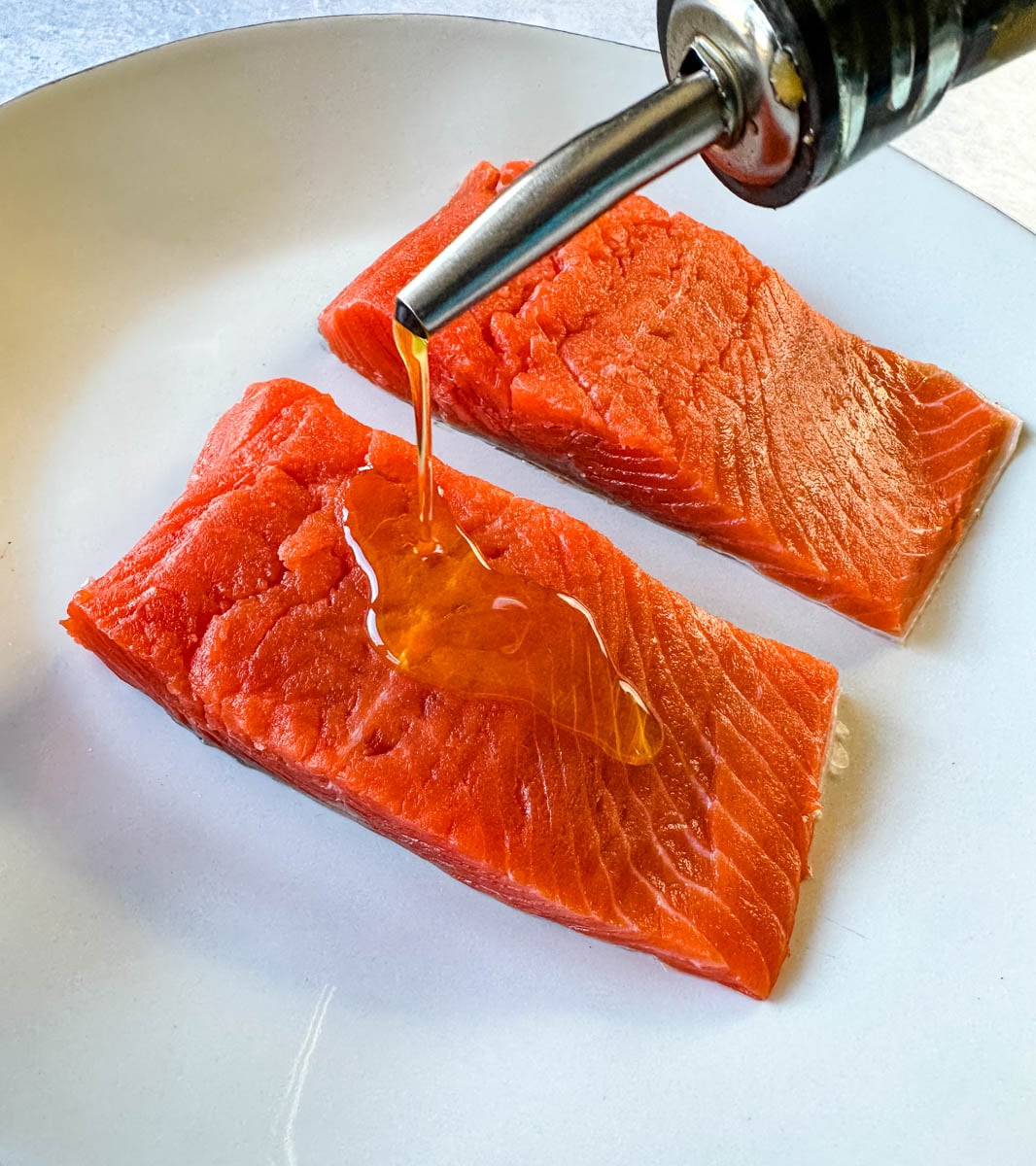 Oil being drizzled over two raw salmon fillets on a white plate.