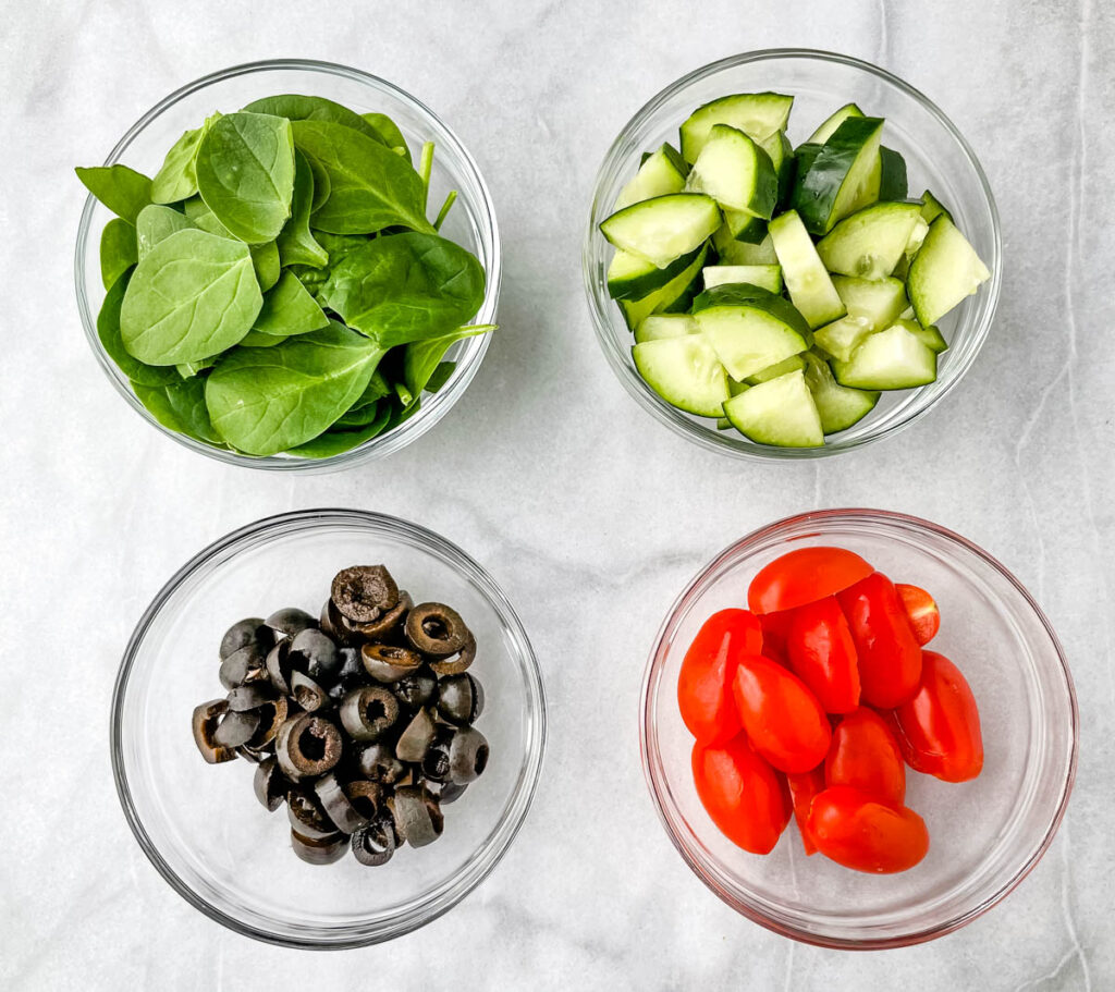 fresh spinach, chopped cucumbers, black olives, and tomatoes in separate glass bowls