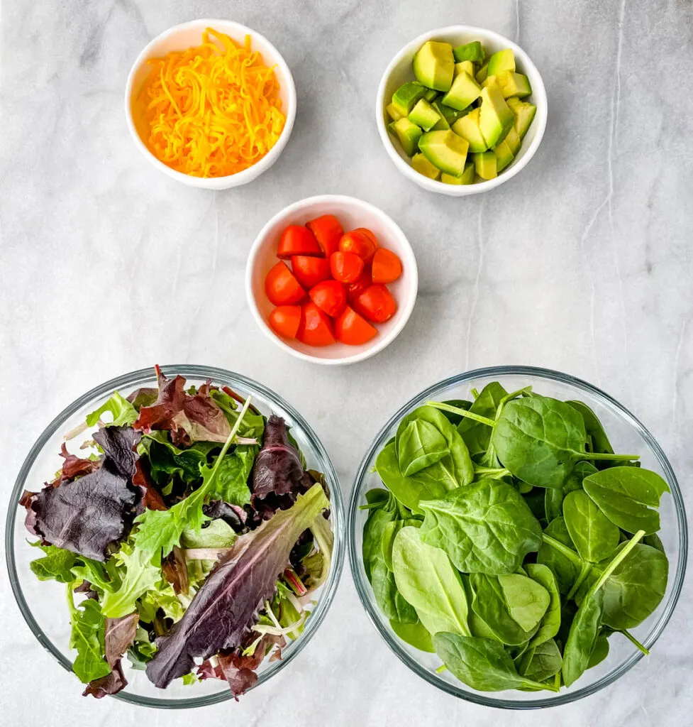 salad greens, spinach, tomatoes, cheddar cheese, avocados, and tomatoes in separate bowls