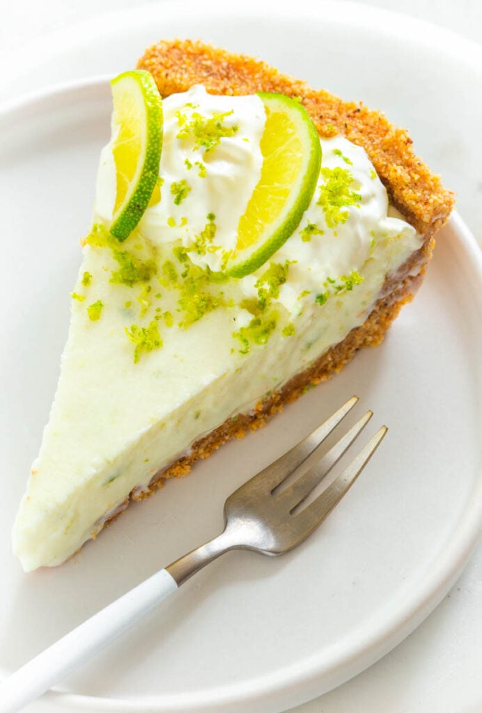 a slice of keto low carb key lime pie on a white plate with fresh limes and a fork