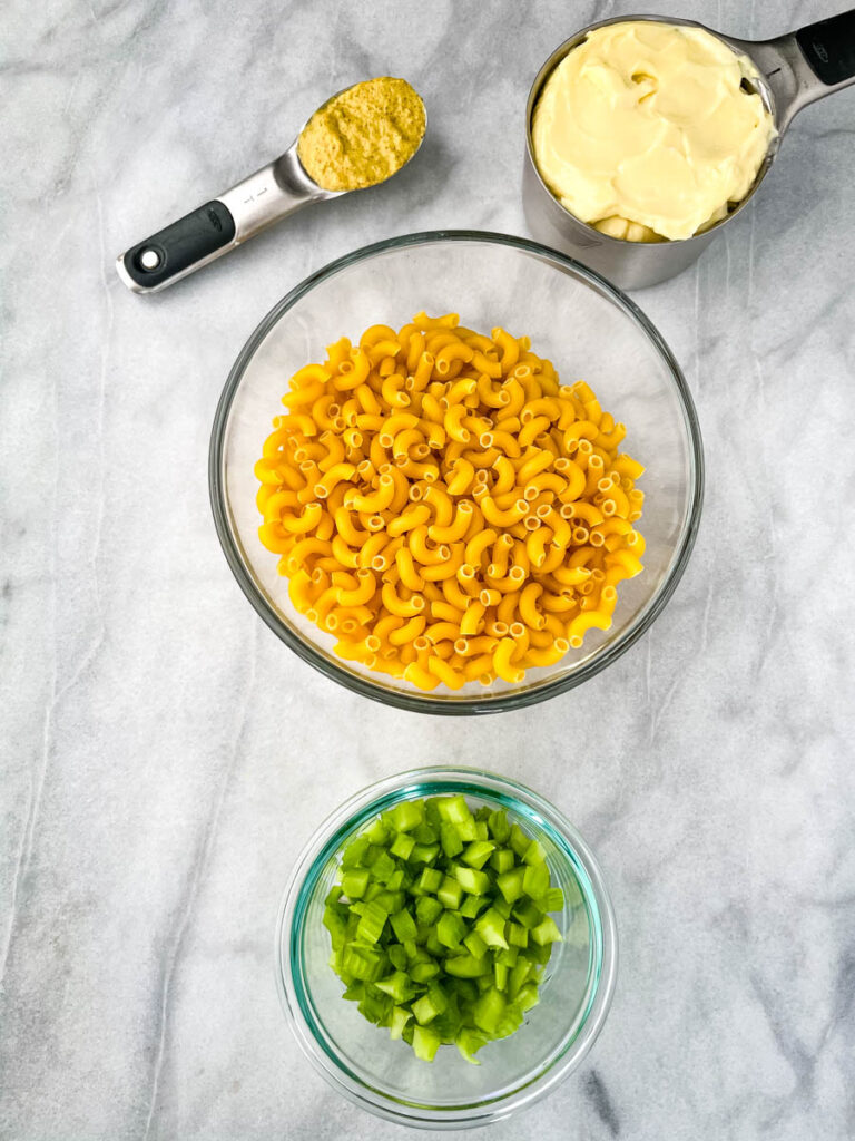 mustard, mayo, uncooked macaroni pasta, and chopped celery in glass bowls