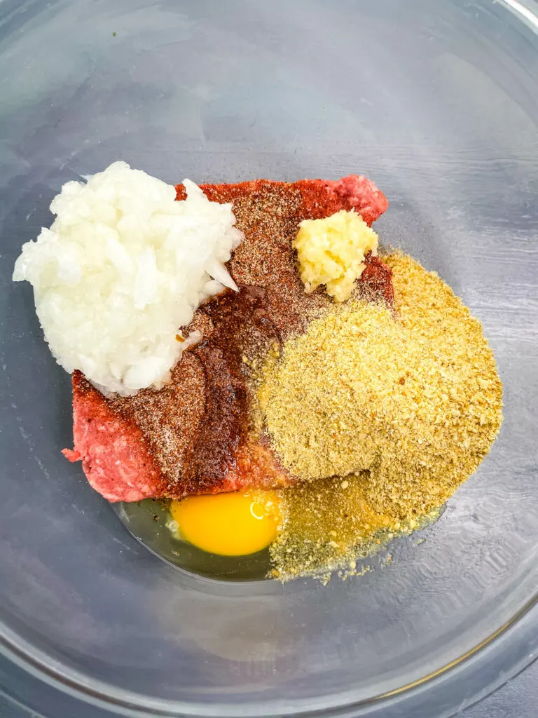 raw ground beef, onions, breadcrumbs, garlic, and an egg in a glass bowl