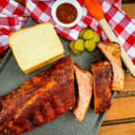 baby back ribs on a plate with bread, BBQ sauce, and pickles