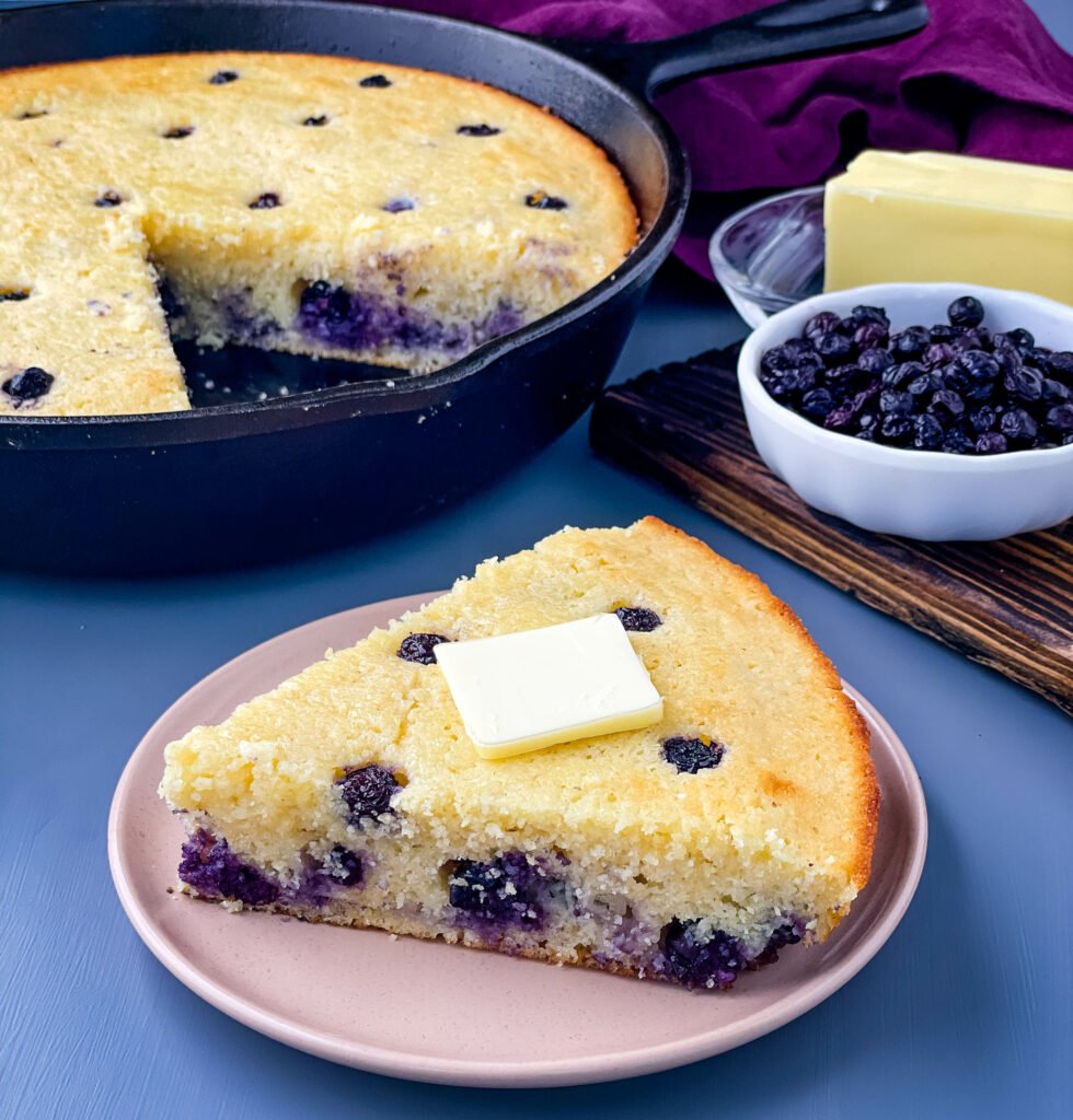 a slice of homemade blueberry cornbread on a plate with a pan of cornbread