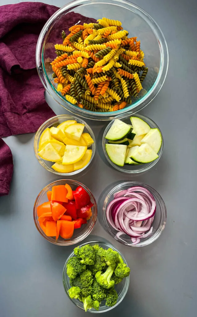 dry rotini pasta, broccoli, red peppers, yellow peppers, green peppers, and red onions in glass bowls