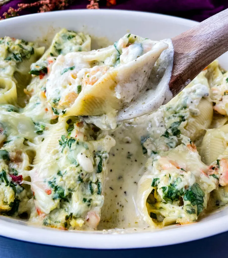 a wooden spoon holding seafood lobster, shrimp, and crab stuffed shells in a baking dish