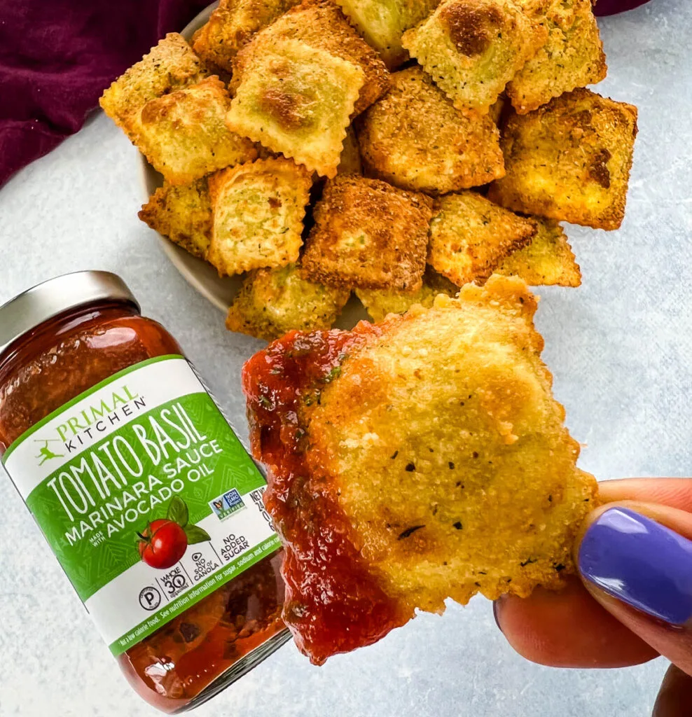 person holding air fryer toasted fried ravioli on a plate with Primal Kitchen marinara sauce