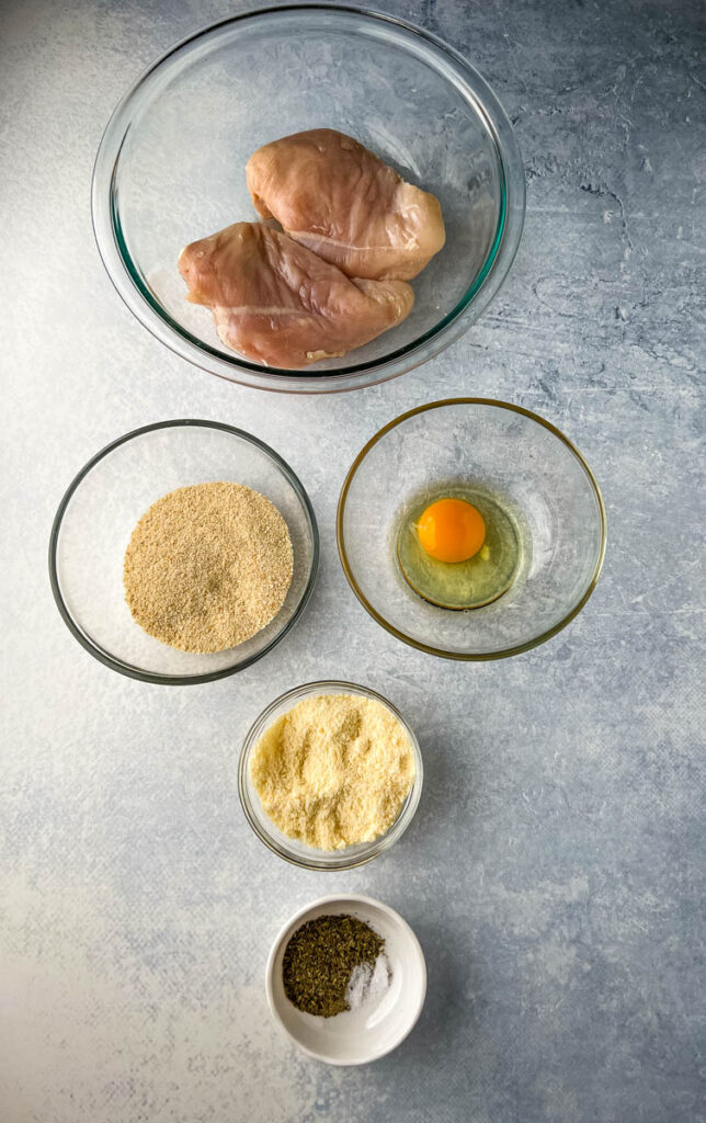 raw chicken breasts, breadcrumbs, eggs, parmesan cheese, and Italian seasoning in separate bowls