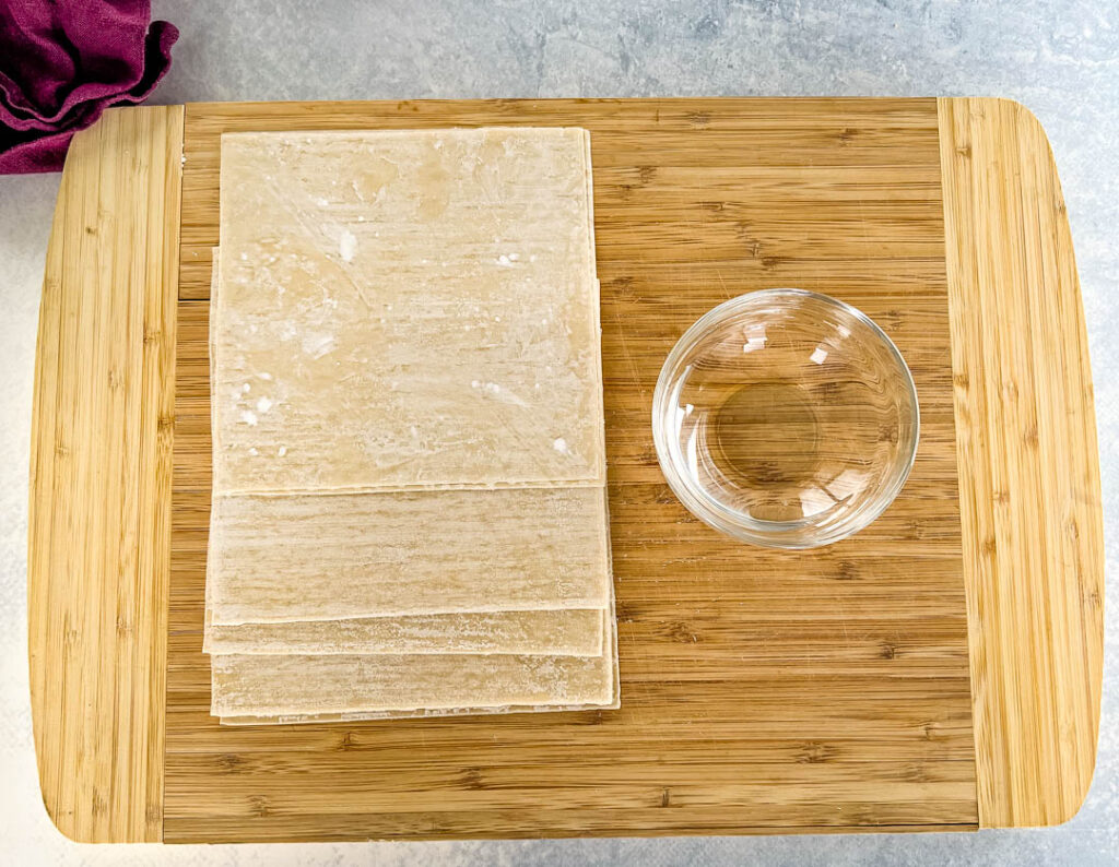 egg roll wrappers and a bowl of water on a bamboo cutting board