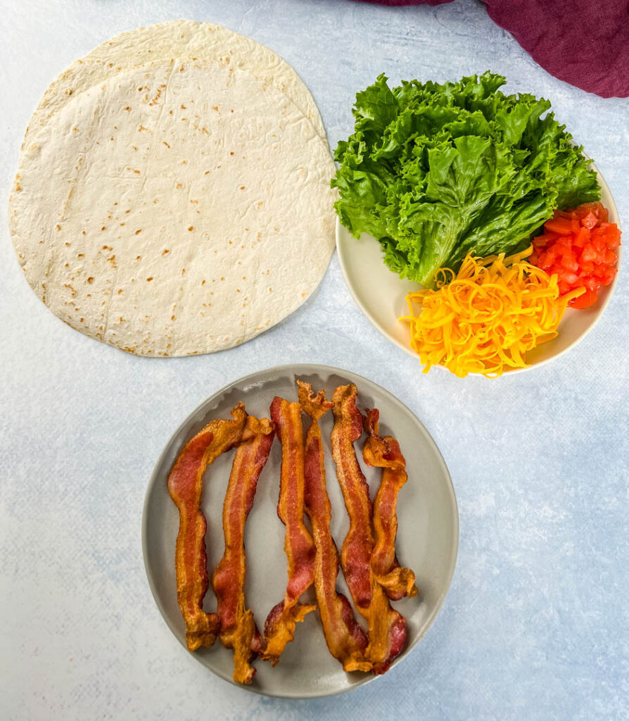 tortilla, cooked bacon, lettuce, tomatoes, and cheese on separate plates