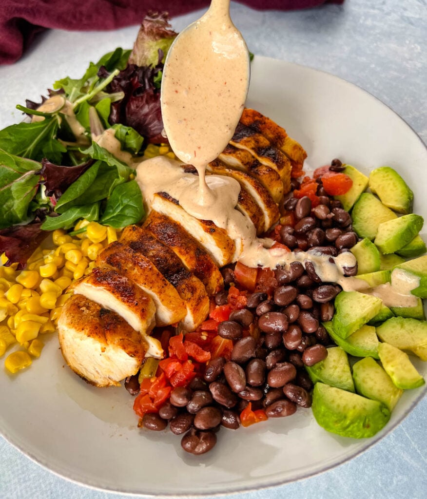 salad dressing drizzled over southwest salad with lettuce, corn, chicken, tomatoes, black beans, and avocado on a plate